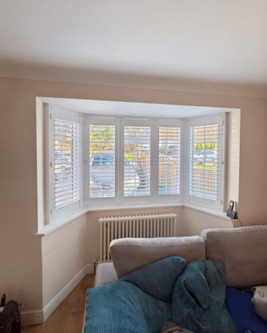 UK Made PVC Plantation Shutters for Angled Bay Living Room Window Beautifully fitted UK made white PVC plantation shutters installed in a living room with an angled bay window by Scarlet Blinds in Manchester, custom made for a perfect fit.